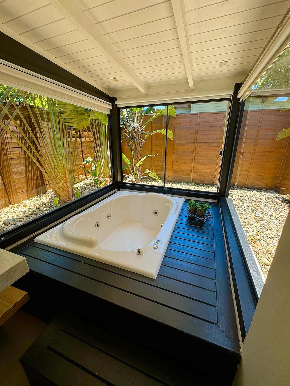 A private jacuzzi is set within a glass enclosure, surrounded by tropical plants. Natural light illuminates the space, highlighting the smooth white surface of the tub and the dark wooden decking underneath.