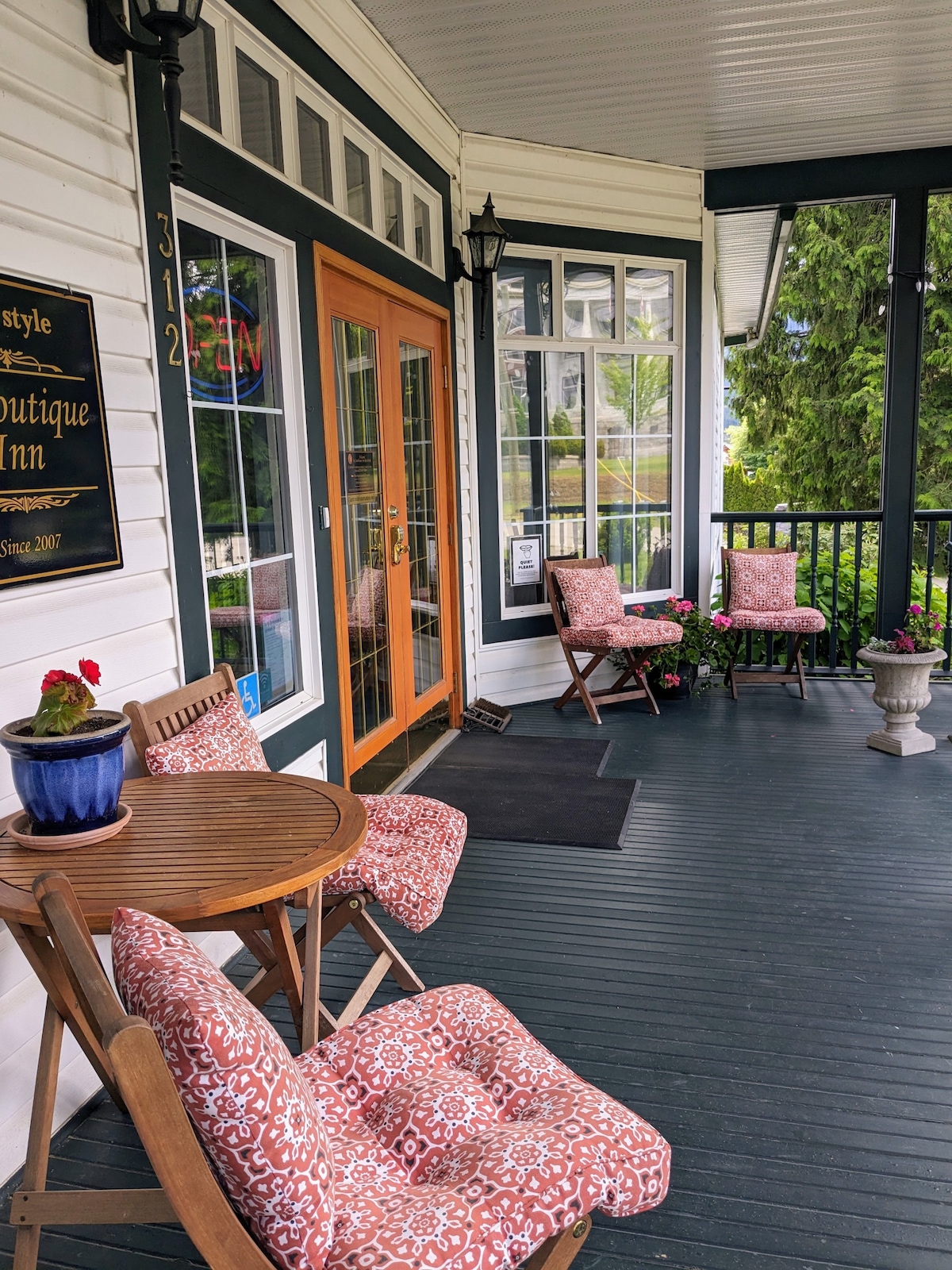 A welcoming front porch features a round table with two folding chairs, each adorned with patterned cushions. Potted flowers add a touch of color, while the entrance door is framed by large windows, allowing natural light to enhance the space.