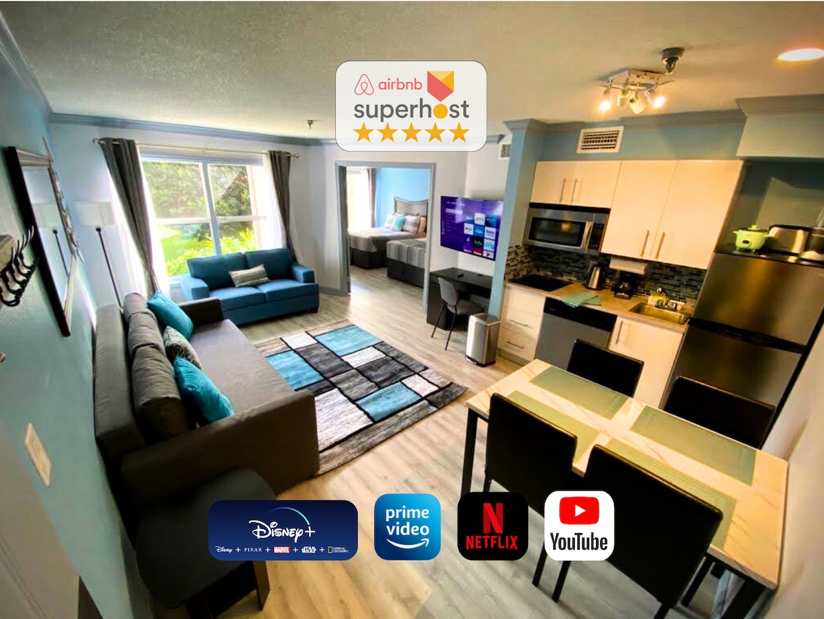 The open living area showcases a comfortable sofa and spacious kitchenette, designed in soft blue tones. Large windows allow natural light to fill the room. A dining table occupies one side, with a modern television mounted on the wall, displaying various streaming service logos.