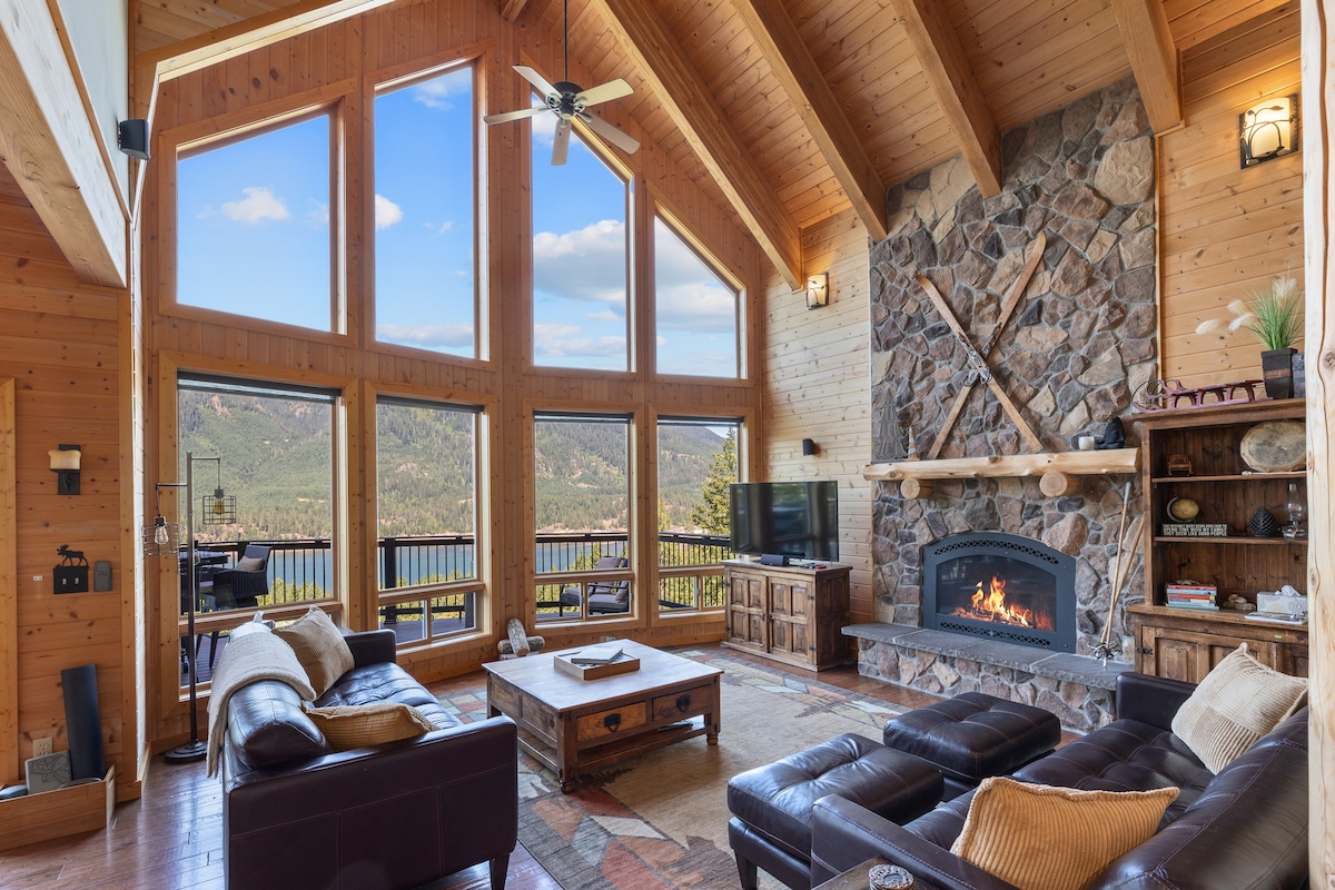 The spacious living area features high wooden ceilings and large windows that frame scenic mountain views. A stone fireplace is situated prominently against one wall, complemented by comfortable leather seating and a wooden coffee table, creating a cozy gathering space.
