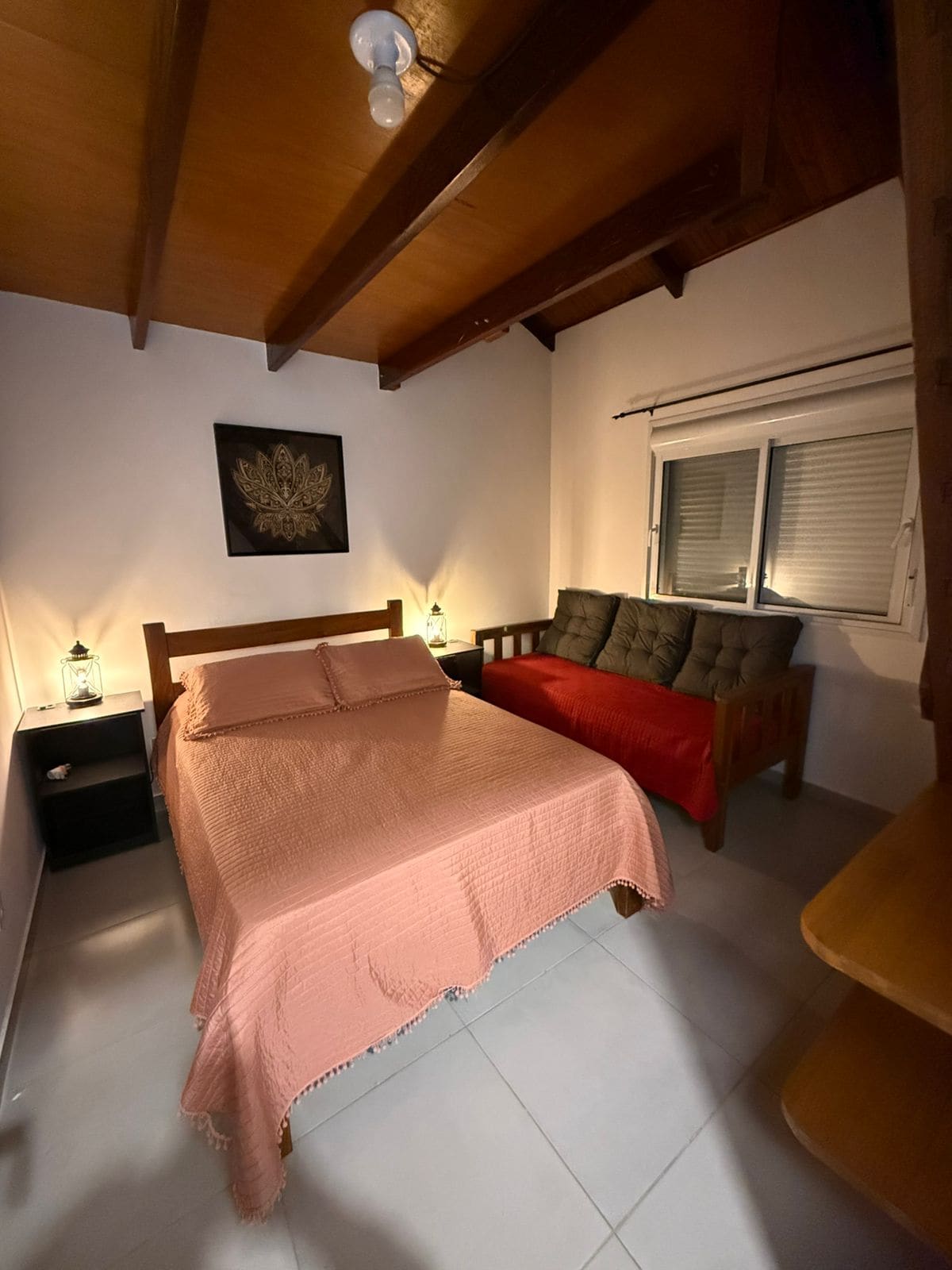 A cozy sleeping area features a spacious bed draped in a light pink coverlet. A comfortable sofa bed is positioned nearby, complemented by subtle bedside lamps. Natural light filters through a partially covered window, and wooden beams add character to the ceiling.