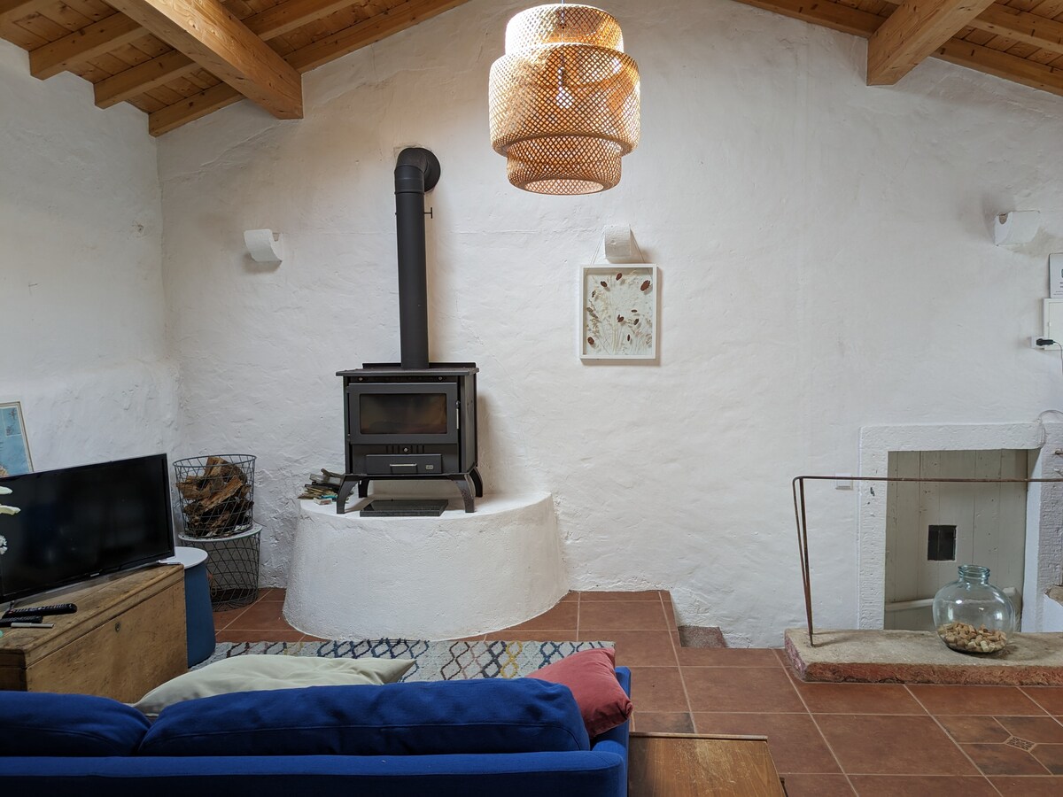 A cozy living area features a blue sofa with cushions, a wood-burning stove set within a white stone structure, and a rustic wooden coffee table. Natural light highlights the textured walls and a woven ceiling lamp hangs above, adding warmth to the space.
