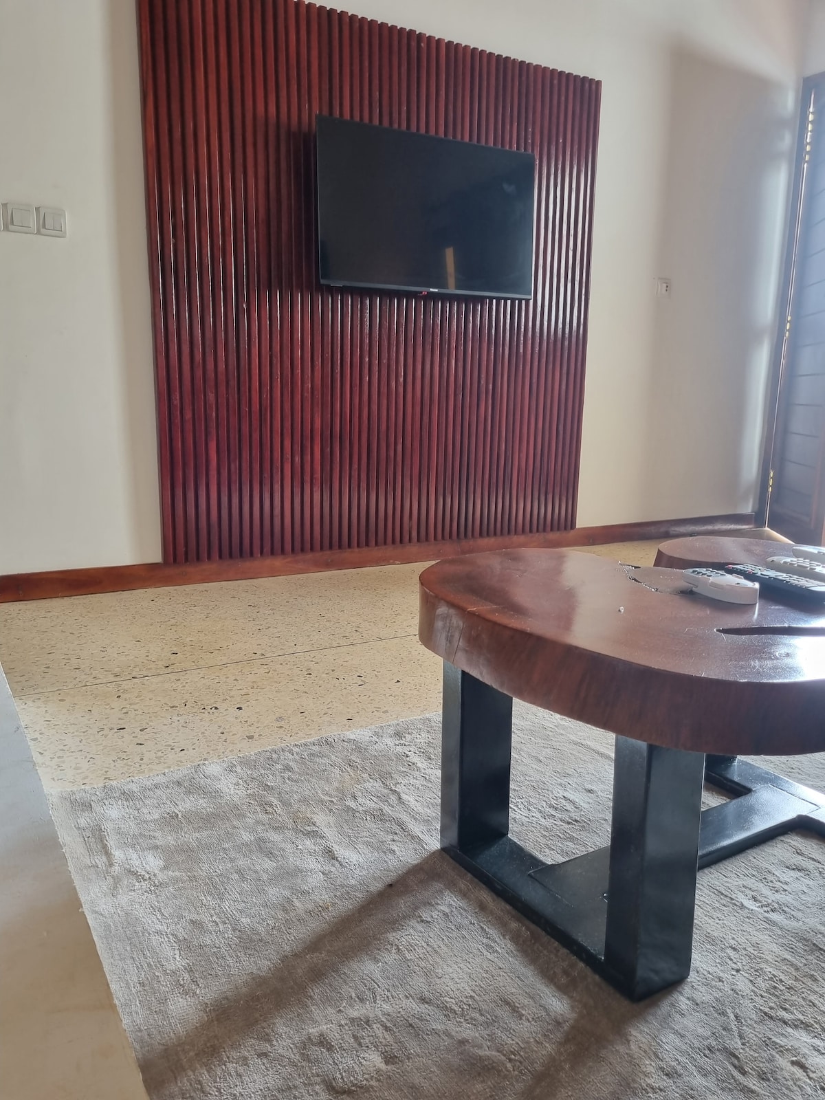 A modern living area features a wooden accent wall with a mounted television. A round coffee table with a dark metal base is positioned over a light-colored rug, adding warmth to the space. Natural light filters through a nearby doorway.