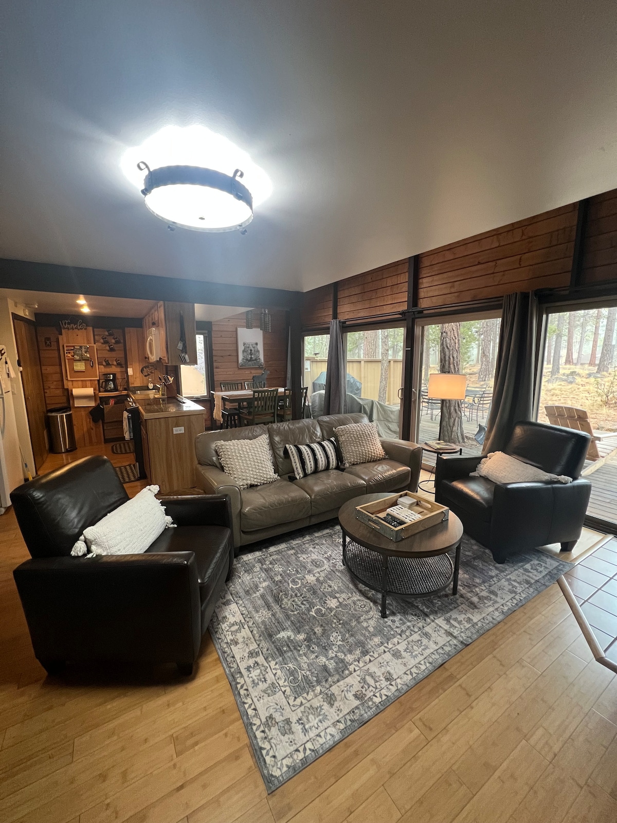 The inviting living area features a comfortable grey sofa and two black armchairs, arranged around a circular coffee table. Natural light flows in through expansive windows, offering views of the pine trees outside. The wood-paneled walls and bamboo flooring create a warm ambiance.