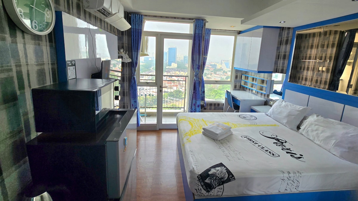 A studio room showcases a spacious bed with white linens and pillows, complemented by a modern kitchenette and mini-fridge. Large windows offer a view of the city, framed by sheer curtains. A clock hangs on the wall, alongside stylish blue accents throughout the space.