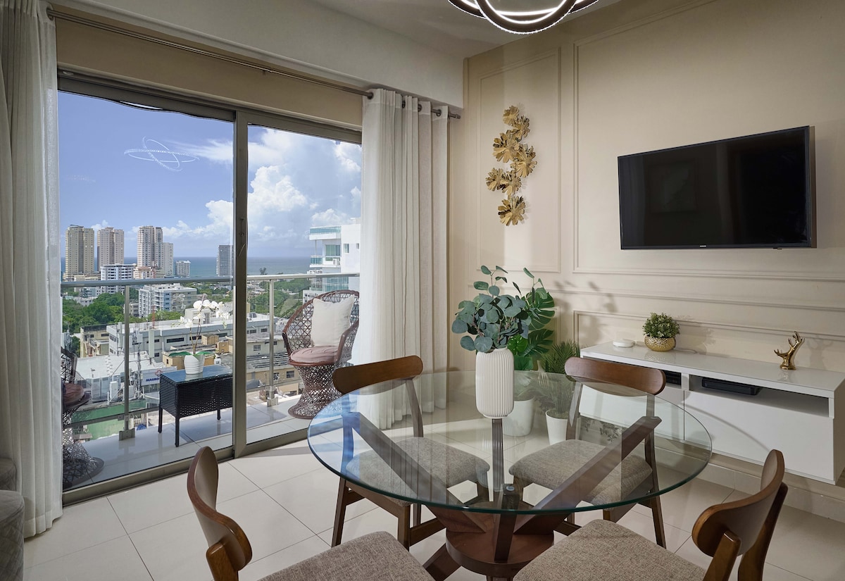 A dining area is presented with a round glass table surrounded by wooden chairs. A large window offers a view of the city and ocean, with curtains drawn to the side. A television is mounted on the wall, complemented by decorative plants and wall art.