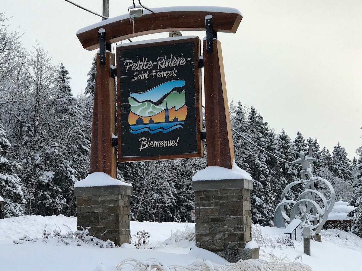 A welcoming sign for Petite-Rivière-Saint-François is showcased, with a depiction of the local landscape. The sign is surrounded by a blanket of snow, and trees dusted with white can be seen in the background, creating a serene winter scene.