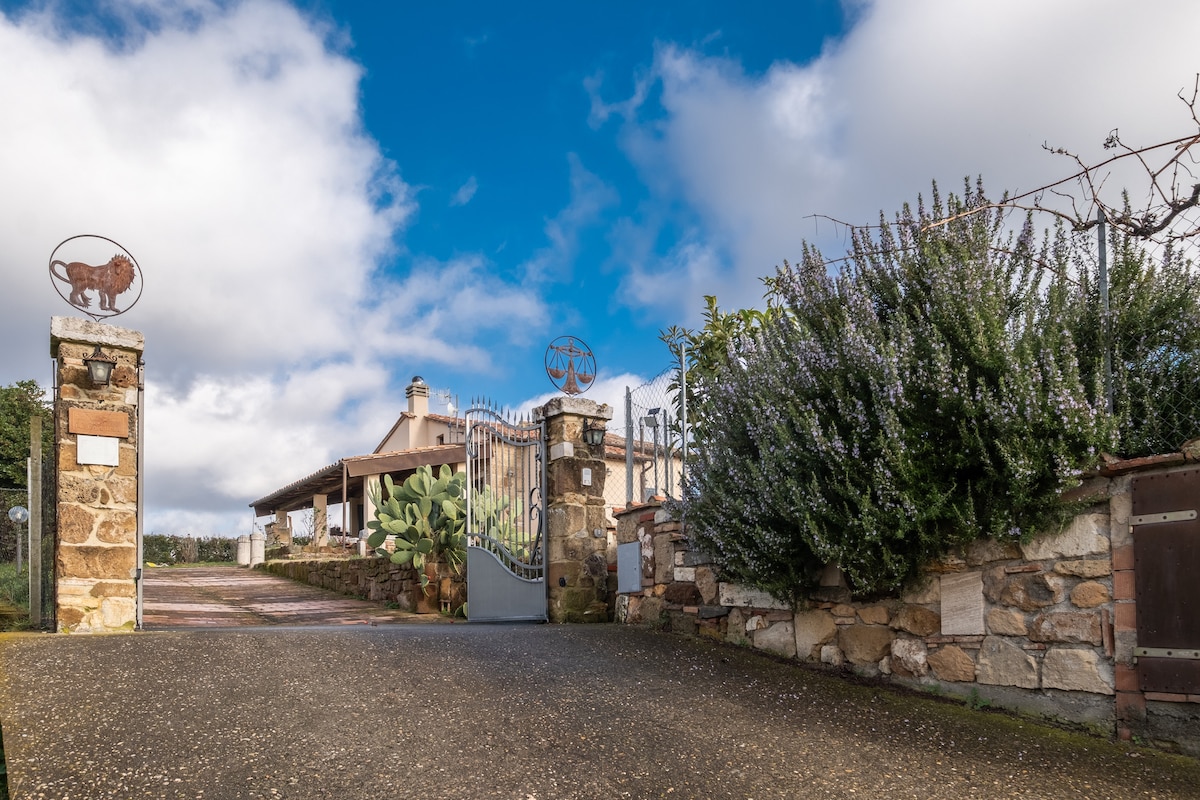 A stone gate marks the entrance to the property, framed by lush greenery and flowering plants. The pathway is lined with low walls, leading to a welcoming covered porch area. A decorative emblem rests atop the gate, enhancing the rustic charm.