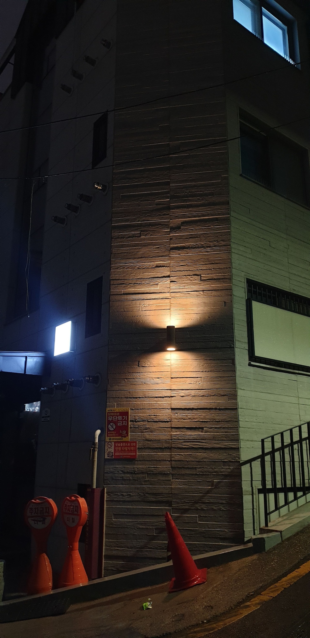 The building's exterior is depicted at night, highlighting its modern architectural features. Soft lighting illuminates the textured stone wall. Two traffic cones are positioned on the side of the pathway, while a window above reflects light, creating a contrast against the darker surroundings.