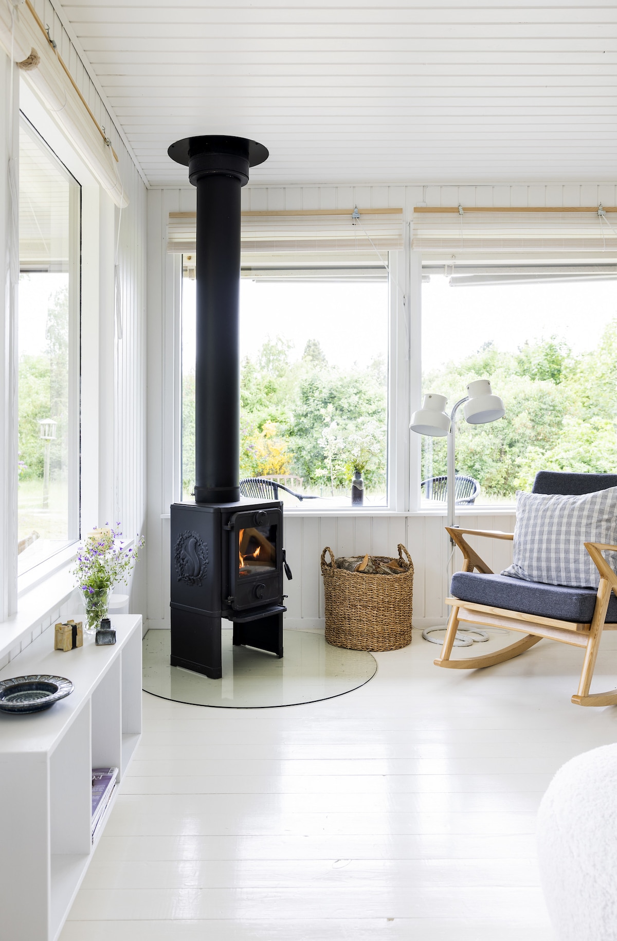 A black wood stove stands elegantly in the corner of a bright room, surrounded by large windows that offer views of lush greenery. A comfortable chair with neutral-toned cushions sits nearby, while a woven basket adds a touch of warmth to the space.