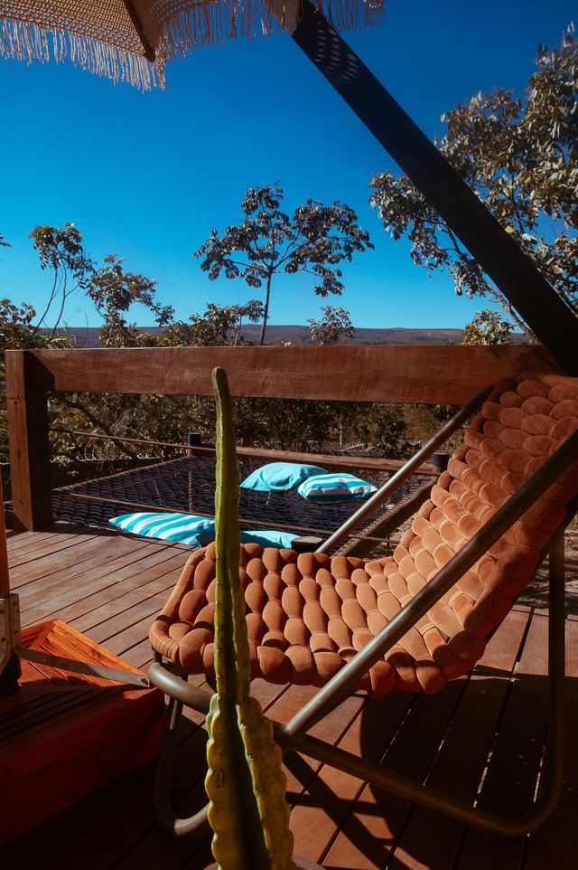 A comfortable hammock chair is positioned on a wooden deck, surrounded by nature. Two blue and white striped loungers are visible in the background, complemented by foliage under a clear blue sky, offering a serene environment for relaxation.