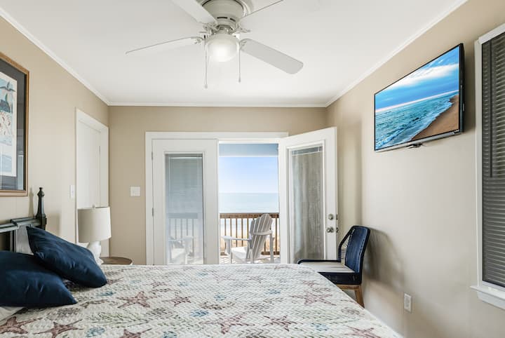 غرفة نوم رئيسية مع مروحة سقف وسرير بحجم كوين وحمام متصل وأبواب مزدوجة تؤدي إلى الشرفة الخاصة المظللة المطلة على المحيط. 