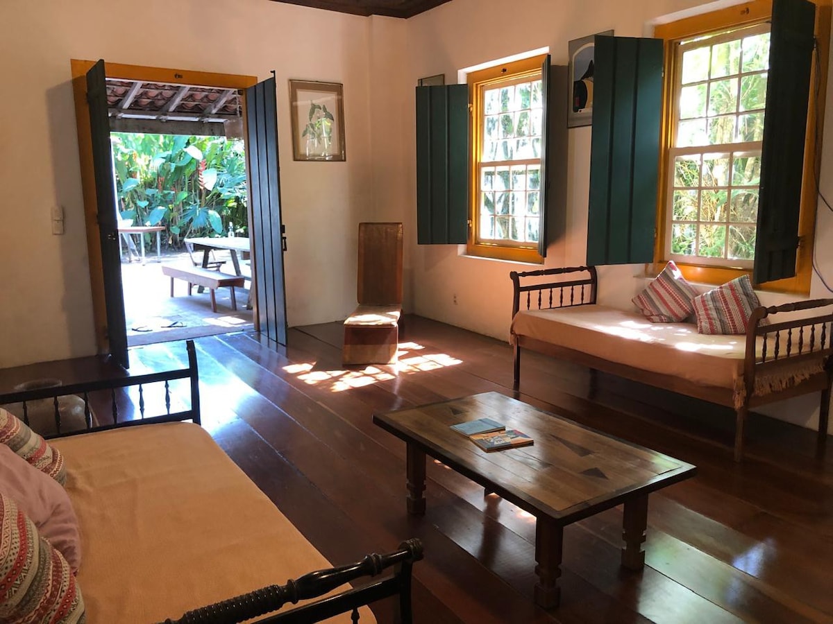 The living area features light wooden flooring and bright yellow window frames, complemented by natural light filtering through opened doors. Two sofas and a wooden chair provide seating options. A wooden table rests at the center, enhancing the communal space, with greenery visible outside.