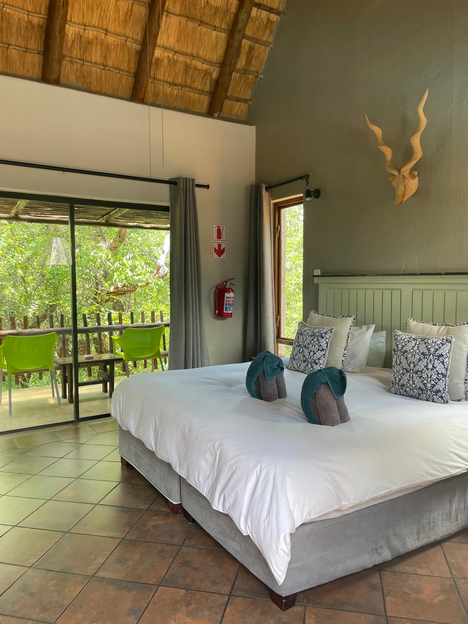 A standard room features a spacious bed with decorative towel arrangements and cushions. A large glass door opens to a private balcony, allowing natural light to illuminate the space. The rustic ceiling and earthy tones complement the surrounding greenery visible outside.