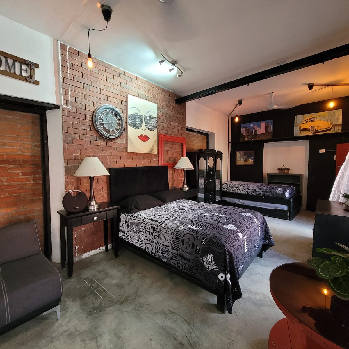 An industrial-style loft is showcased, featuring a cozy seating area with a single chair and a decorative wall with exposed brick. A wall clock and two lamps flank a dark-colored bed with patterned bedding. The space is illuminated by overhead lights, enhancing the ambiance.