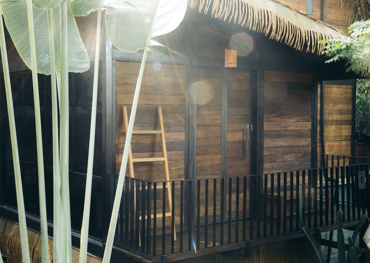 A wooden cabin features a thatched roof and sliding glass doors, offering a glimpse of the outdoor deck. A wooden ladder leans against the cabin's side, while tall green plants frame the entrance, enhancing the natural setting.