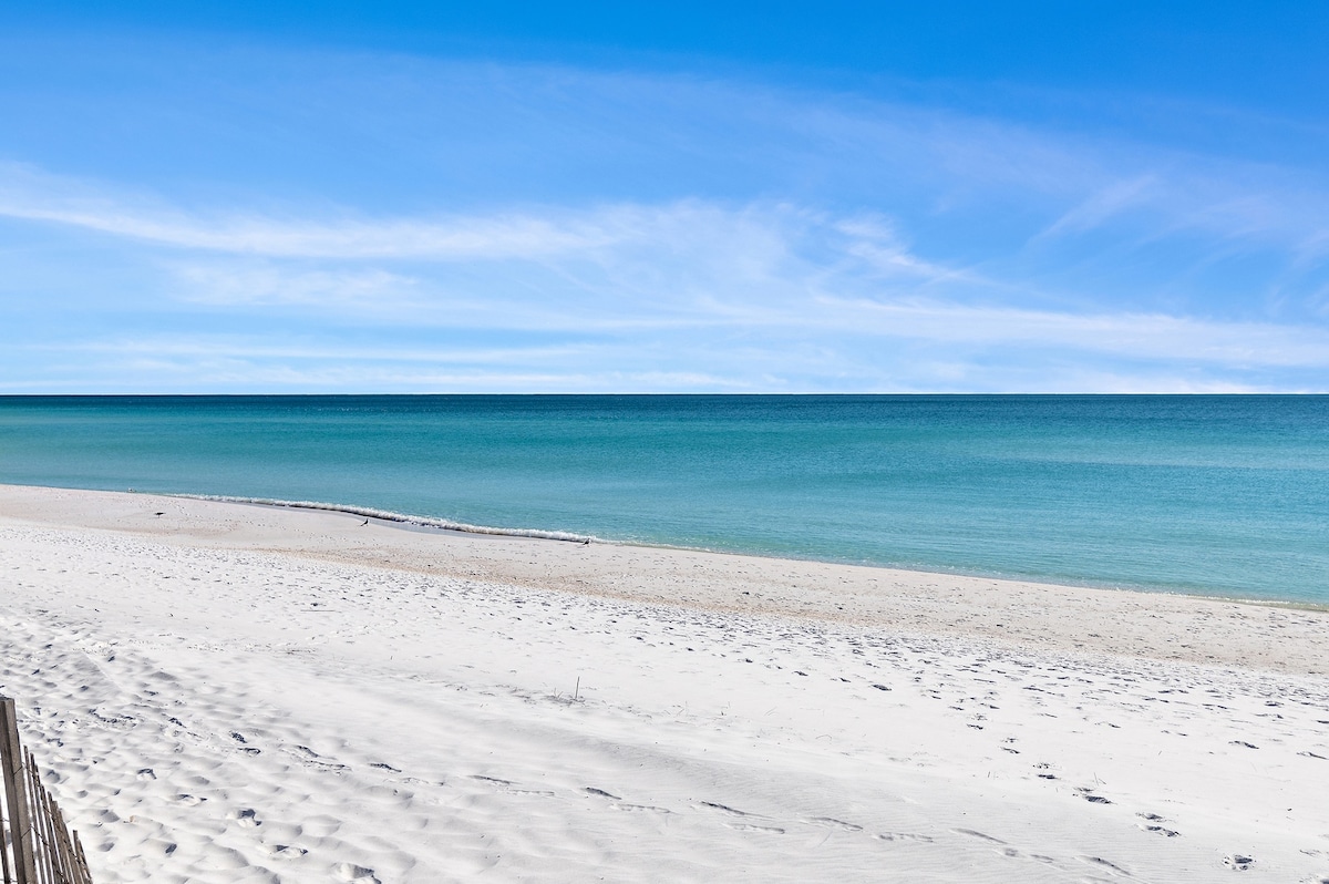 A serene beachfront view presents soft white sands meeting a clear turquoise ocean. The horizon stretches across the image, showcasing gentle waves lapping against the shore. A clear blue sky enhances the tranquil ambiance, inviting relaxation and enjoyment of the natural surroundings.