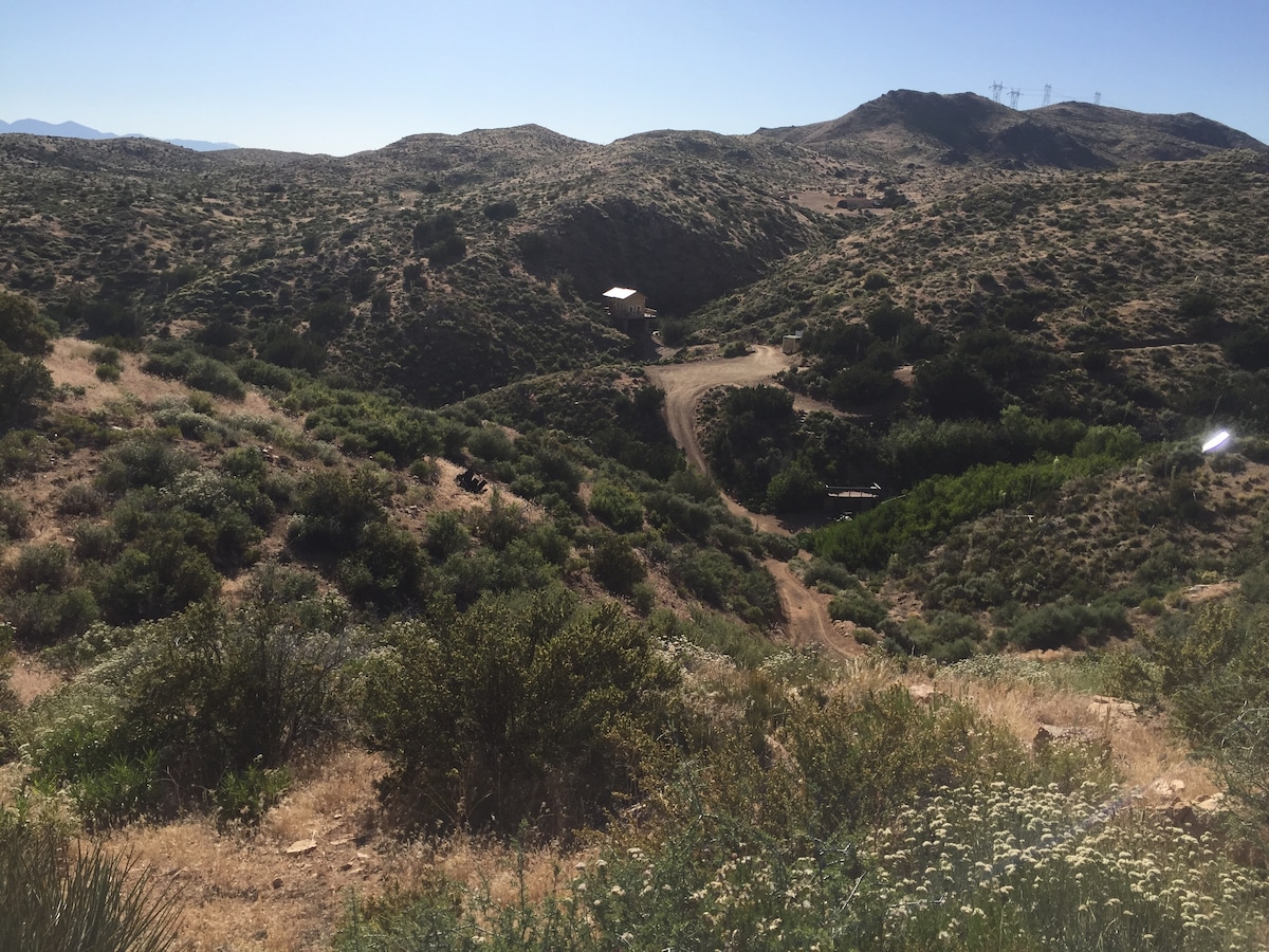 An expansive view of rolling hills blanketed in various shades of green and brown is presented, showcasing a natural landscape. A dirt path winds through the terrain, leading to a distant structure, while the clear blue sky provides a vibrant backdrop.