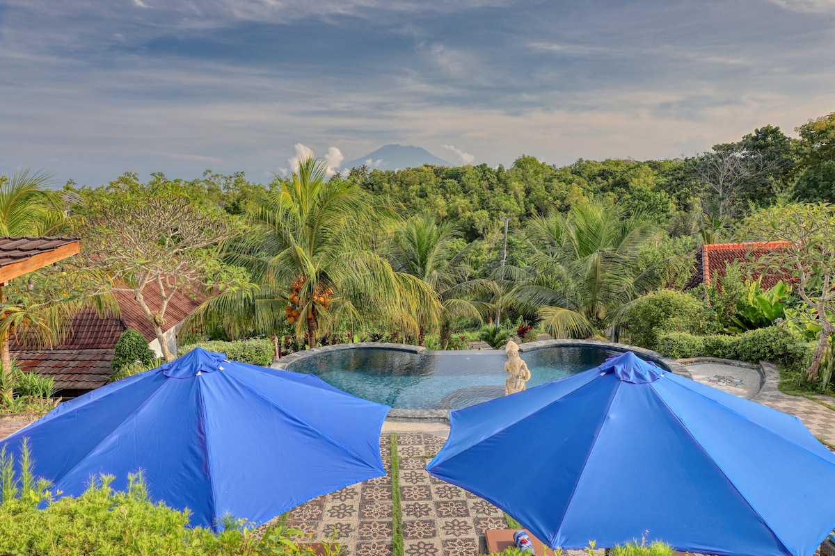 A shared swimming pool is framed by tropical greenery and hills, with Mount Agung visible in the distance. Two large blue umbrellas offer shade beside the pool, with patterned stone flooring and lush plants enhancing the serene outdoor setting.