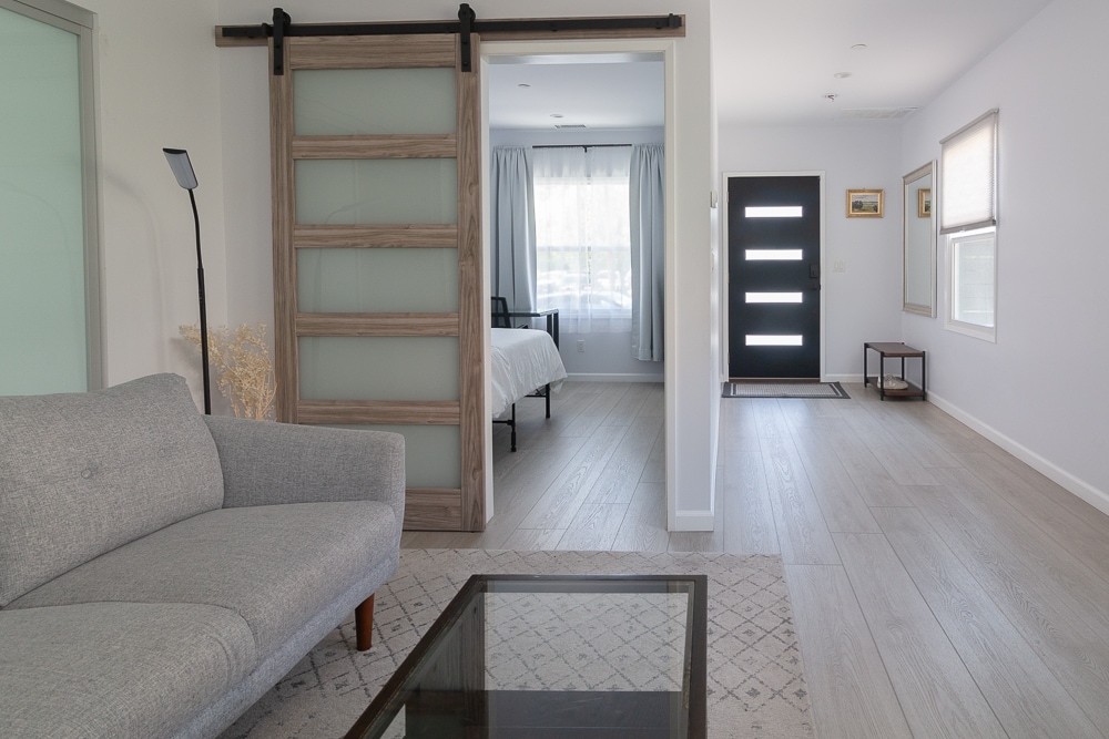 A modern living area showcases a light gray couch facing a glass coffee table. A sliding barn door leads to the bedroom, while the entryway features a large front door and bright natural light filtering through sheer curtains.