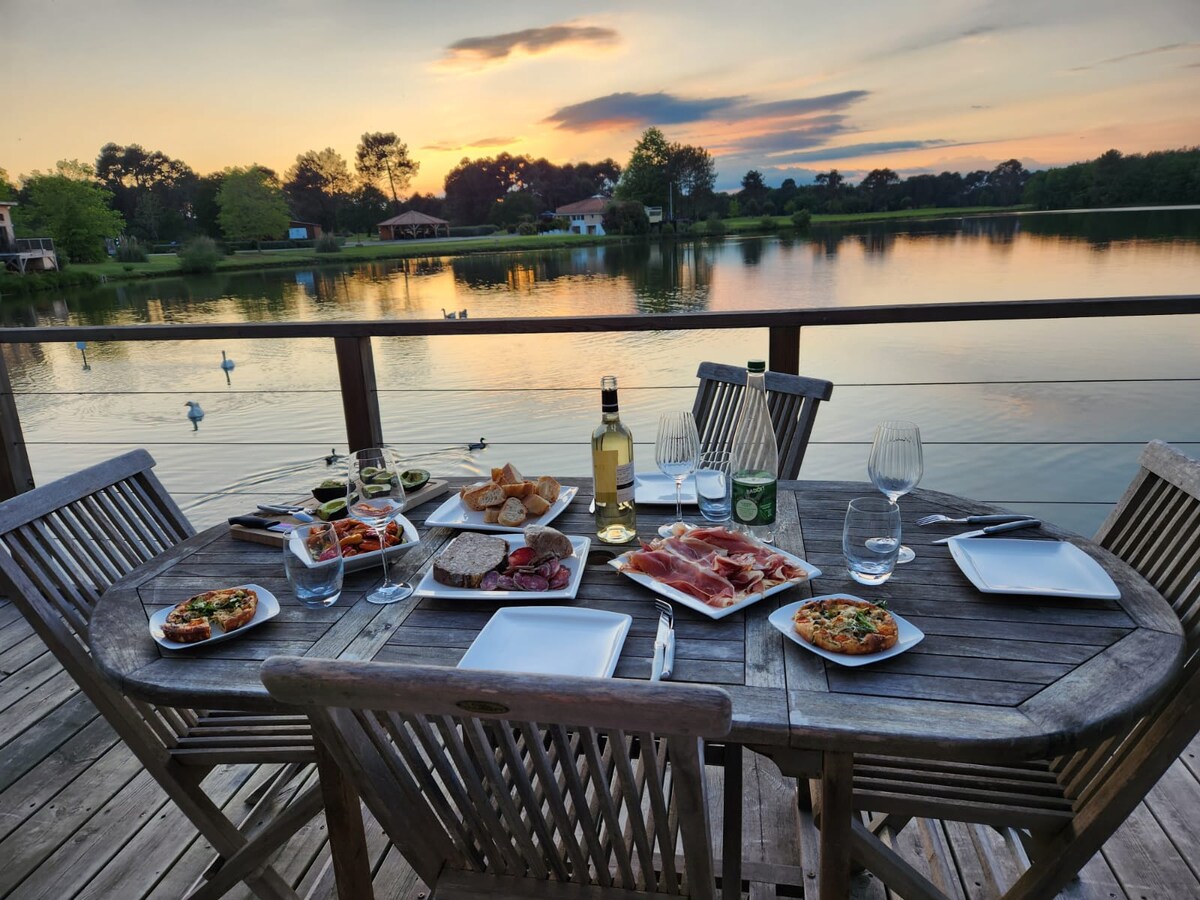 A wooden dining table is set for a meal, featuring several plates of food and glasses. A bottle of wine stands on the table, with a serene lake and lush greenery in the background, illuminated by the soft hues of the sunset.