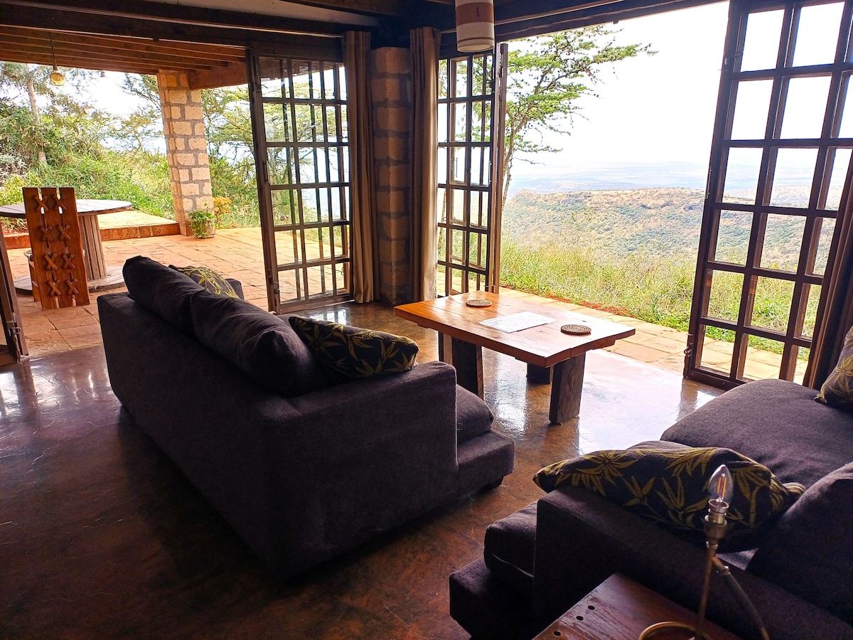 The indoor living area features comfortable grey sofas with patterned cushions, positioned near large glass doors that open to an expansive outdoor patio. A wooden coffee table sits in the center, while the lush greenery and scenic views of the valley are visible through the open doors.