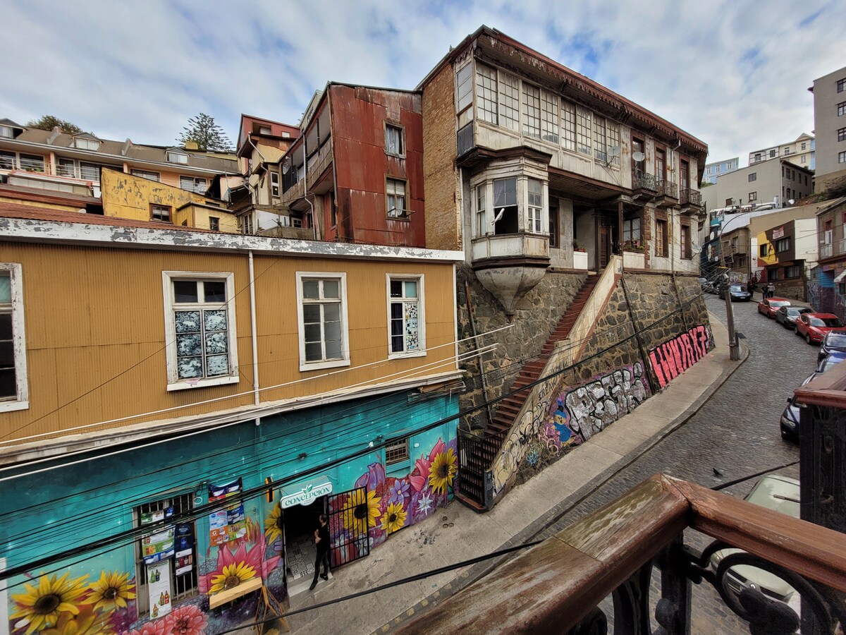 The image depicts a vibrant street scene featuring a multi-story building with a weathered facade and a contrasting brightly painted mural on the adjacent wall. The street is lined with art and flowers, while steps lead up to the entrance of the prominent building.