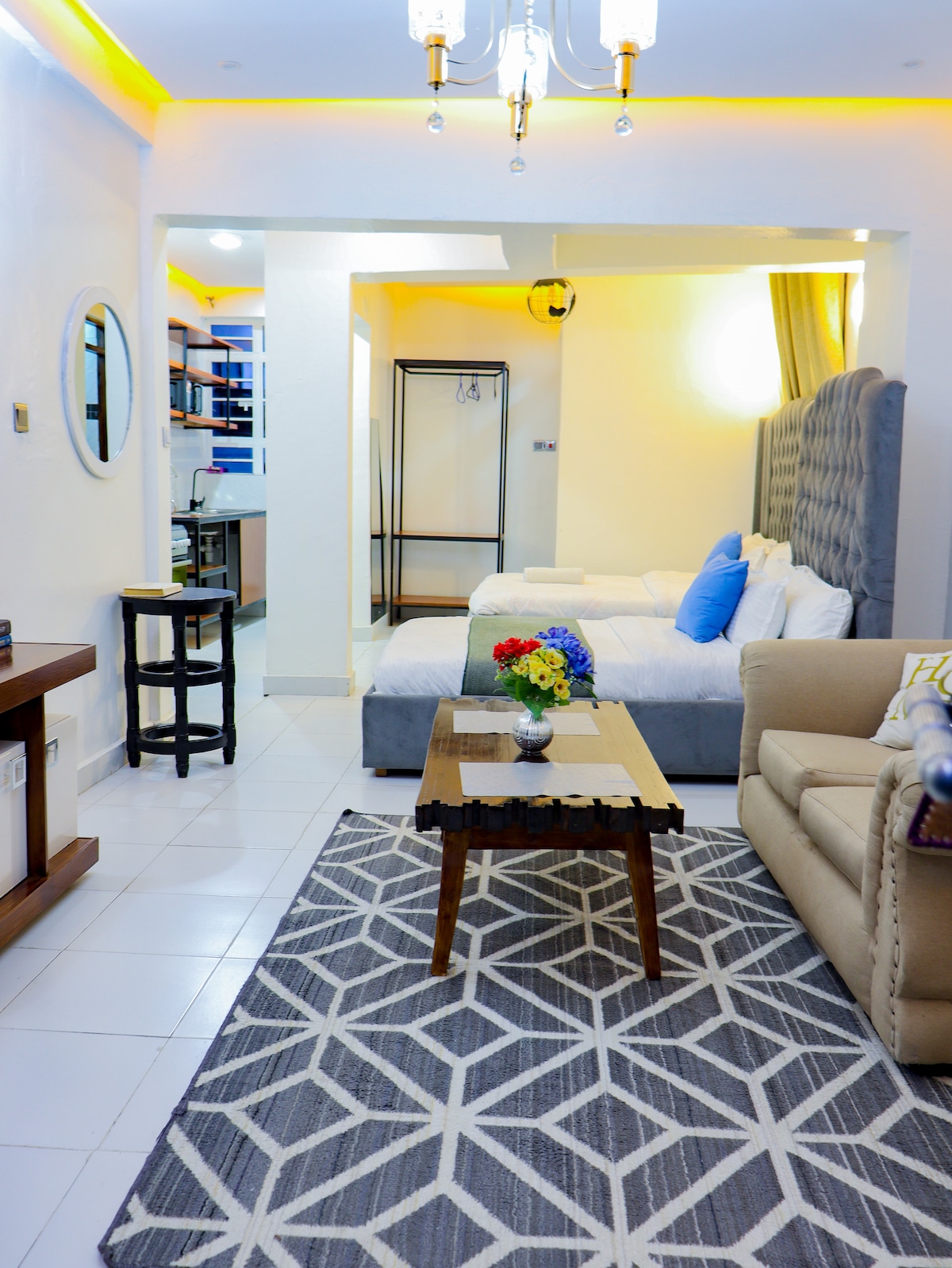 A well-lit living area features a comfortable sofa and a center table, adorned with a vase of flowers. In the background, a spacious bedroom is visible with two beds, complemented by soft bedding. A geometric patterned rug enhances the inviting ambiance.