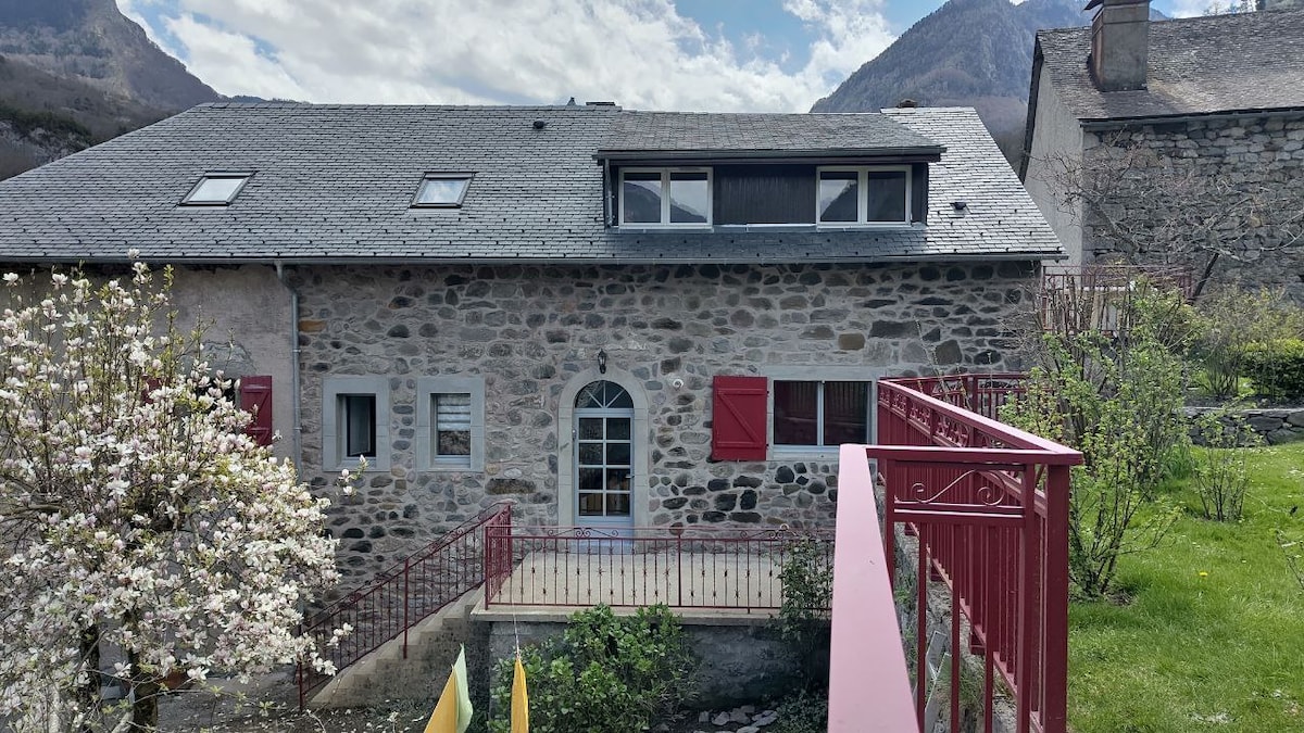 The exterior of a stone building is shown, featuring a balcony with a red railing. Large windows with shutters highlight the entrance, which is framed by a small wooden deck. A flowering tree and grassy area are visible in the foreground, with mountains in the background.