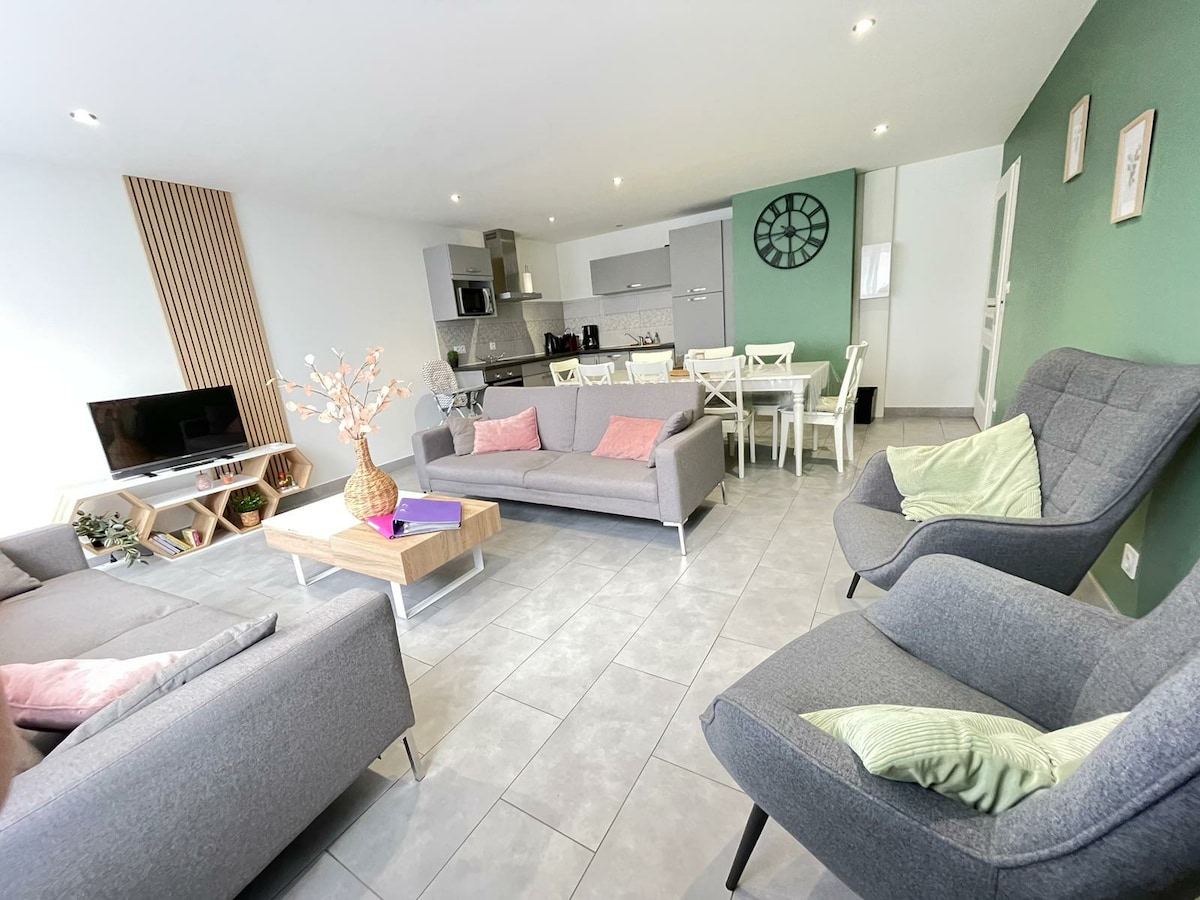 A modern living area is presented, featuring two spacious sofas and two stylish armchairs. A light wood coffee table sits at the center. The open-plan space includes a dining area with a table, surrounded by white chairs. The kitchen is visible, showcasing sleek appliances and cabinetry.