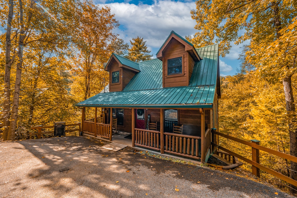 A rustic cabin is set against a backdrop of vibrant autumn foliage, showcasing a green metal roof and two gabled sections with dark framed windows. A welcoming front porch features multiple rocking chairs, inviting relaxation in the natural surroundings.