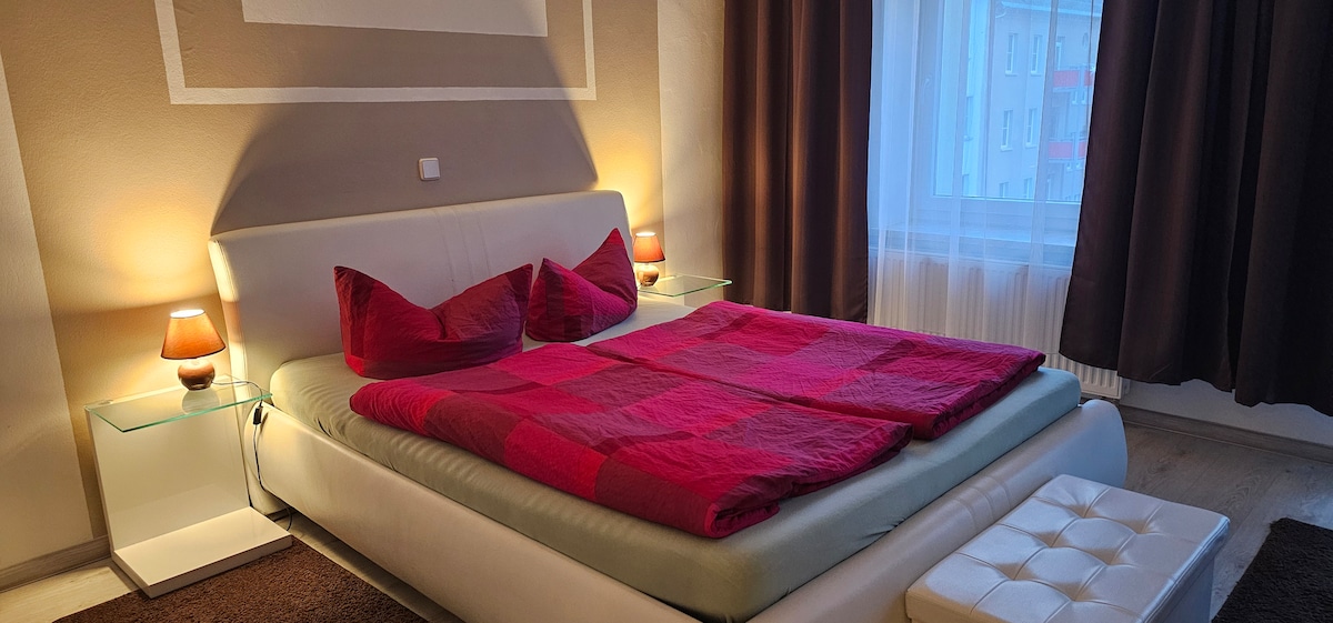 The bedroom features a spacious bed dressed in a vibrant red blanket. Nightstands with soft lamps sit on either side, providing gentle lighting. Plush curtains frame the window, allowing natural light to enter, while a small ottoman is placed at the foot of the bed.