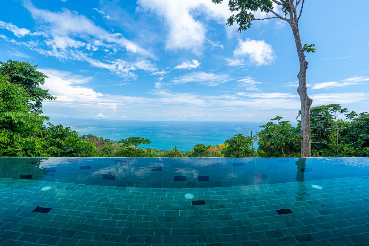 An infinity edge saltwater pool mirrors the sky, providing uninterrupted views of the Pacific Ocean and surrounding jungle. The clear blue water contrasts beautifully with lush greenery, creating a serene atmosphere. Bright blue skies and scattered clouds add to the inviting setting.