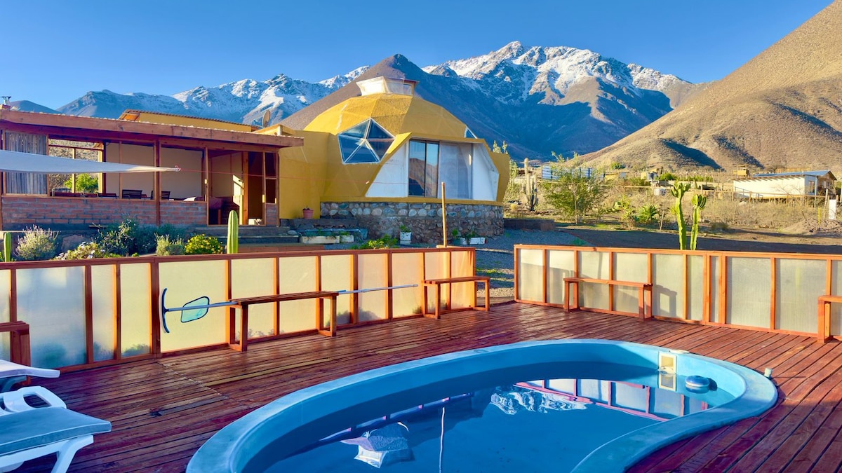 A hexagonal structure is nestled against a mountainous backdrop, with snow-capped peaks visible in the distance. A wooden deck surrounds a small pool, and lounge chairs are arranged nearby, inviting relaxation under clear skies.