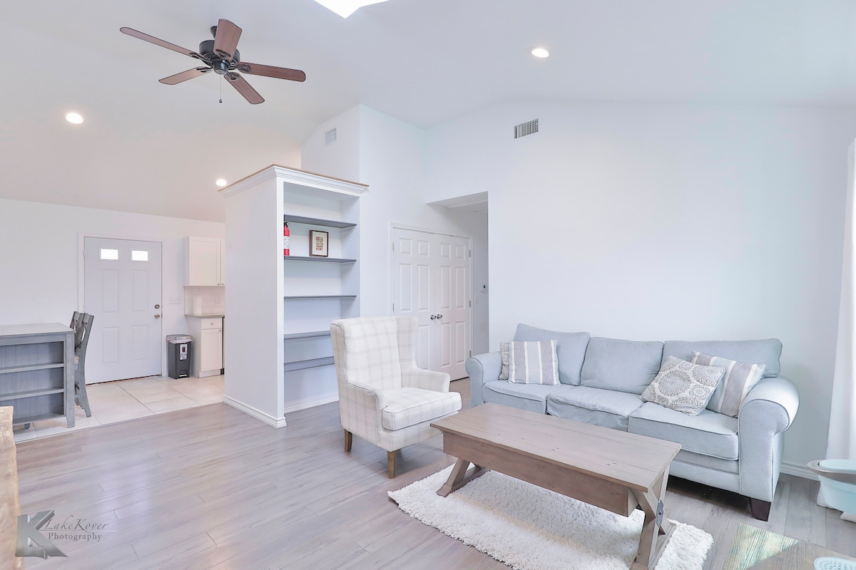A comfortable living area features a light gray sofa and a cozy armchair, positioned around a wooden coffee table. Natural light fills the space through a nearby window, highlighting the neutral color scheme and clean lines of the furnishings.