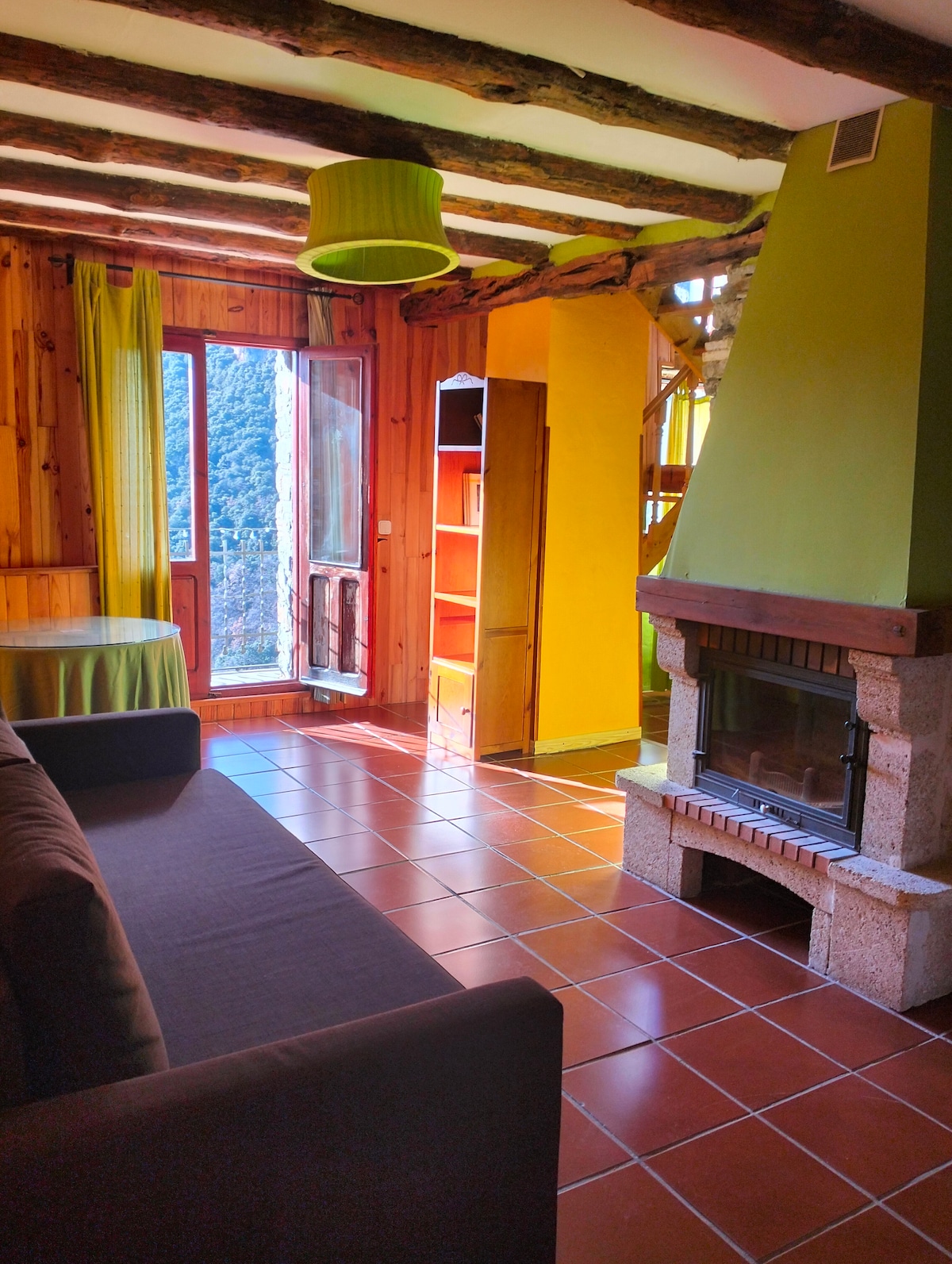 A living area features a stone fireplace set in a green chimney. Sunlight filters through a window, highlighting wooden walls and a circular table. The space includes a dark sofa and a tall shelving unit in the background, contributing to a cozy ambiance.