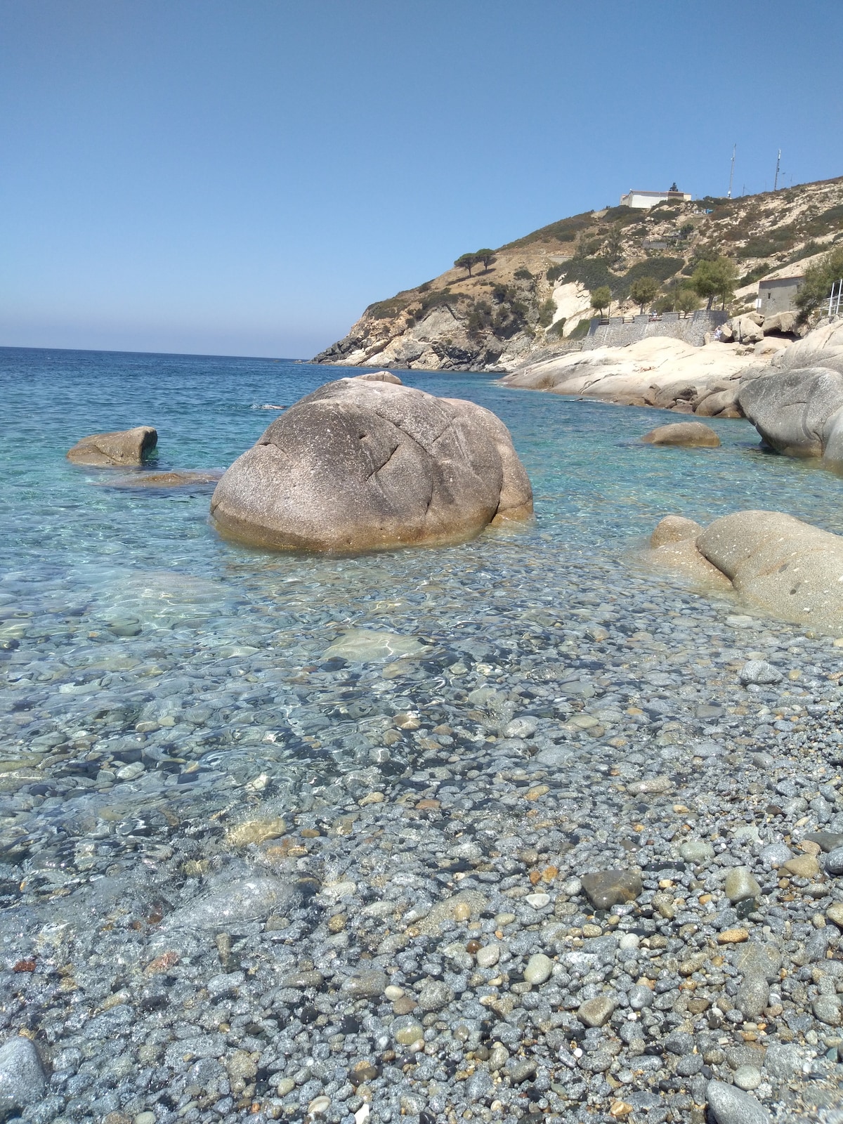 A clear coastal scene features smooth stones and pebbles lining the shoreline. Gentle waves lap against a large rock partially submerged in the water. The tranquil blue sea extends towards a hillside covered in greenery, providing a serene backdrop under a clear sky.