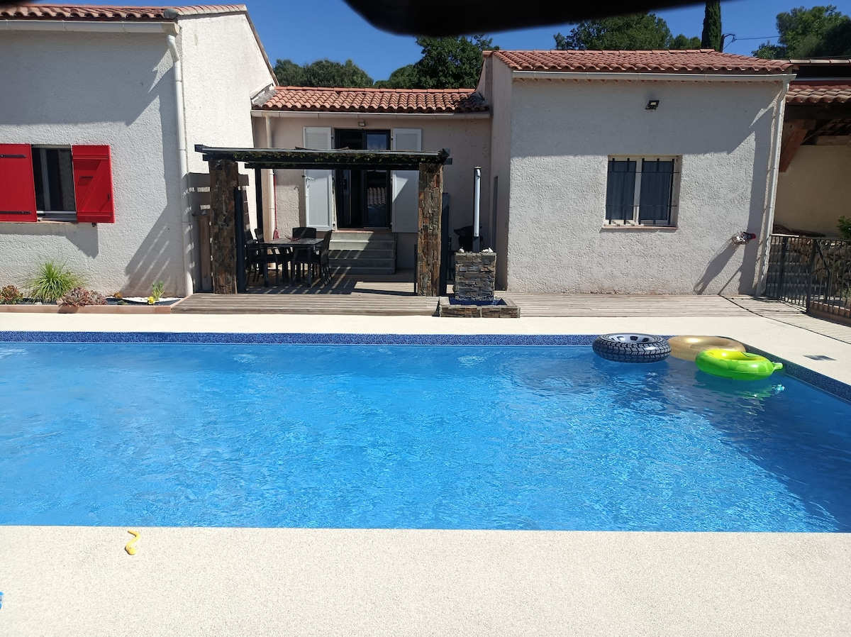 A clear blue swimming pool reflects sunlight, bordered by a paved area. In the background, a partially shaded terrace with a dining table is observed, adjacent to the house's exterior features. Colorful pool floats are positioned on the water's surface, adding a playful touch to the setting.