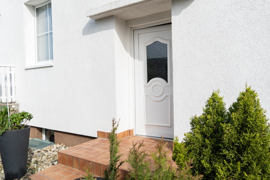 An entrance door framed by a light-colored wall showcases a decorative design. Steps lead up to the door from a tiled pathway, while neatly trimmed shrubs and planters are positioned alongside, enhancing the welcoming appearance of the entryway.