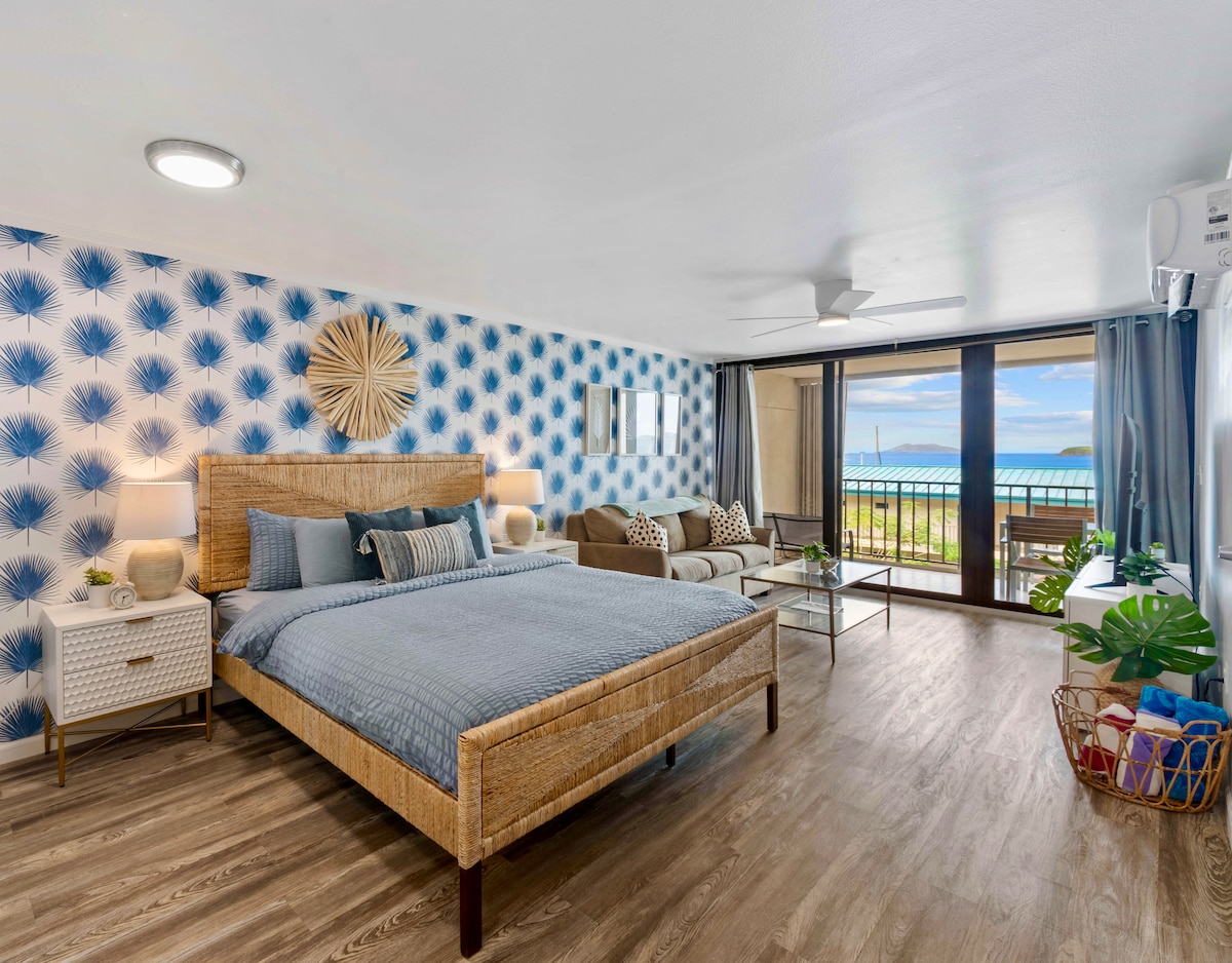 A spacious bedroom is furnished with a large bed featuring a woven headboard. Light blue and white palm-patterned wallpaper adds a serene touch. A seating area with two chairs and a coffee table is visible, and sliding glass doors lead to a balcony overlooking the beach.
