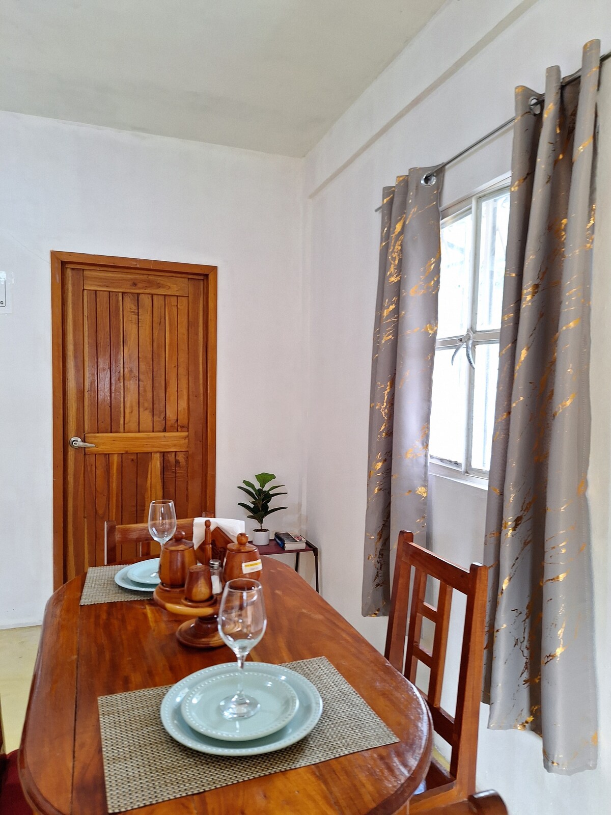 A dining area is highlighted, featuring a wooden table set for two with plates and glasses. Natural light enters through a window dressed with light curtains. A wooden door leads to another room, and a small plant adds a touch of greenery.