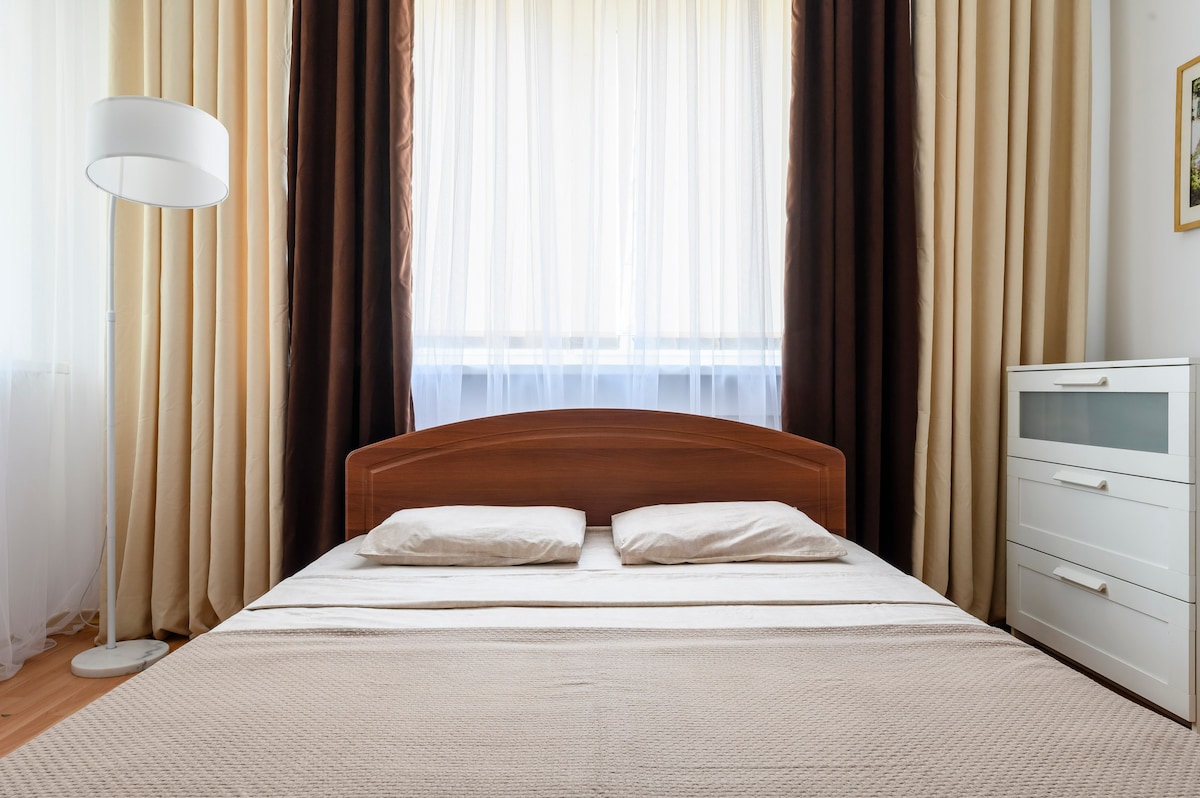 A spacious sleeping area features a wooden bed frame with two soft pillows positioned on a neatly arranged bedspread. Soft curtains in light and dark tones frame a large window, while a modern floor lamp and a dresser with horizontal drawers complement the room's design.