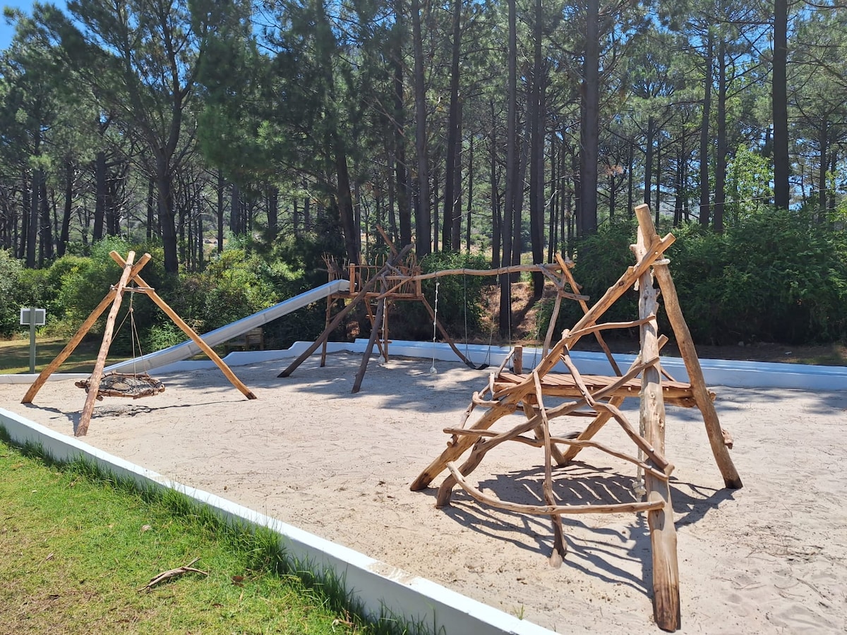 A nature-inspired playground features wooden structures arranged on a sandy area. The playground is surrounded by tall trees, creating a natural setting. The equipment includes large climbing frames and swings, designed to blend harmoniously with the outdoor environment.