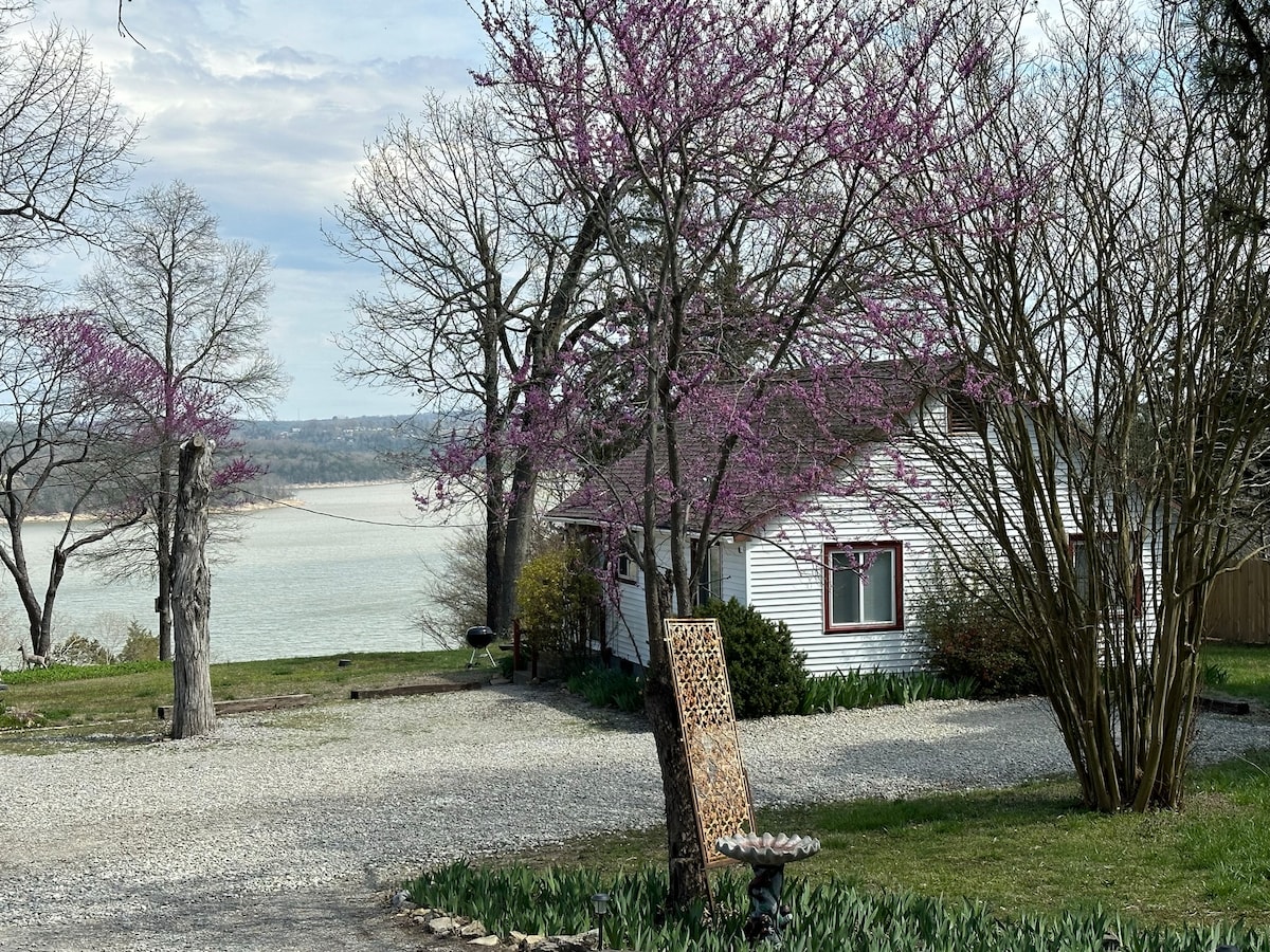 A scenic view of the cabin is framed by blooming purple trees and a gravel driveway. The lake is visible in the background under a cloudy sky, while lush green plants surround the cabin, adding a touch of nature to the setting.