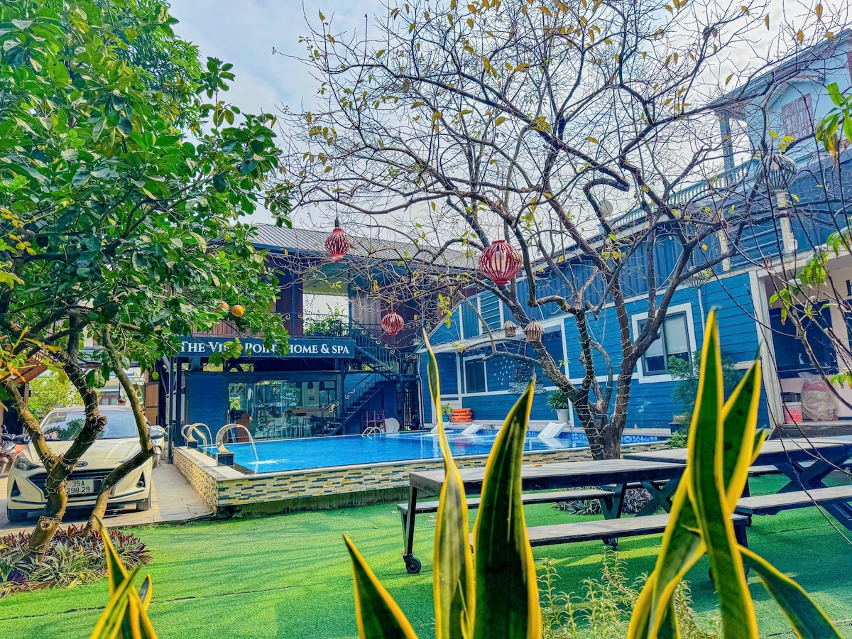 A welcoming outdoor area features a swimming pool with clear blue water surrounded by lush green lawn. Nearby, vibrant plants and trees provide natural shade. A charming building displays the name 'The Viet Pool Home & Spa,' with lanterns hanging from branches, adding a festive touch.