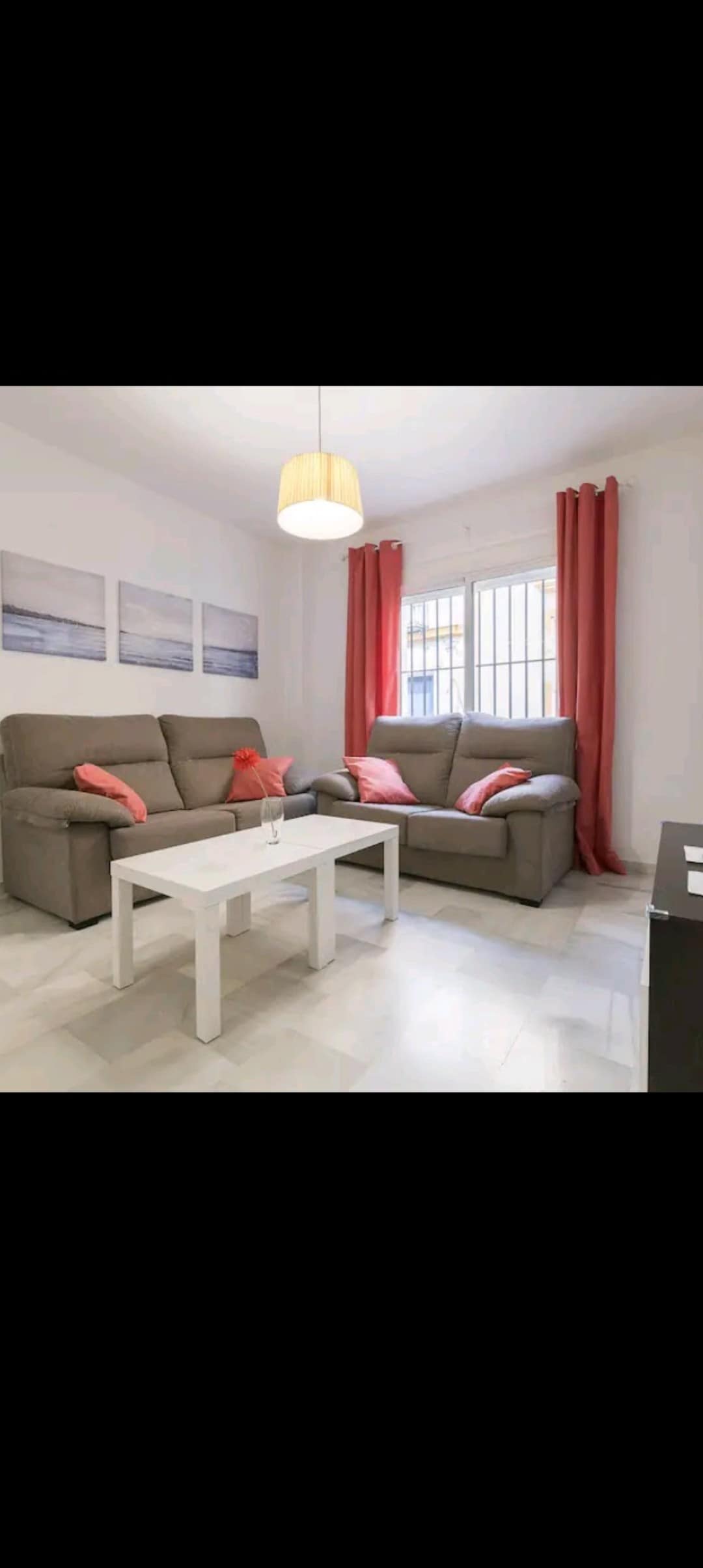 A comfortable living area is arranged with two grey sofas adorned with coral cushions. A white coffee table sits in the center, complementing the light-colored tile flooring. Natural light enters through a window lined with white curtains, while minimal artwork adorns the walls.