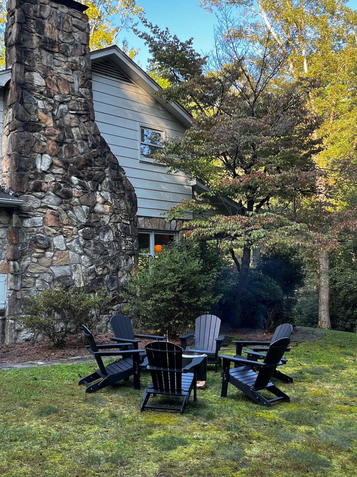 An outdoor seating area features six Adirondack chairs arranged in a circular formation around a fire pit. The stone chimney of the house stands prominently in the background, partially hidden by lush greenery and trees, creating a serene environment for gatherings.
