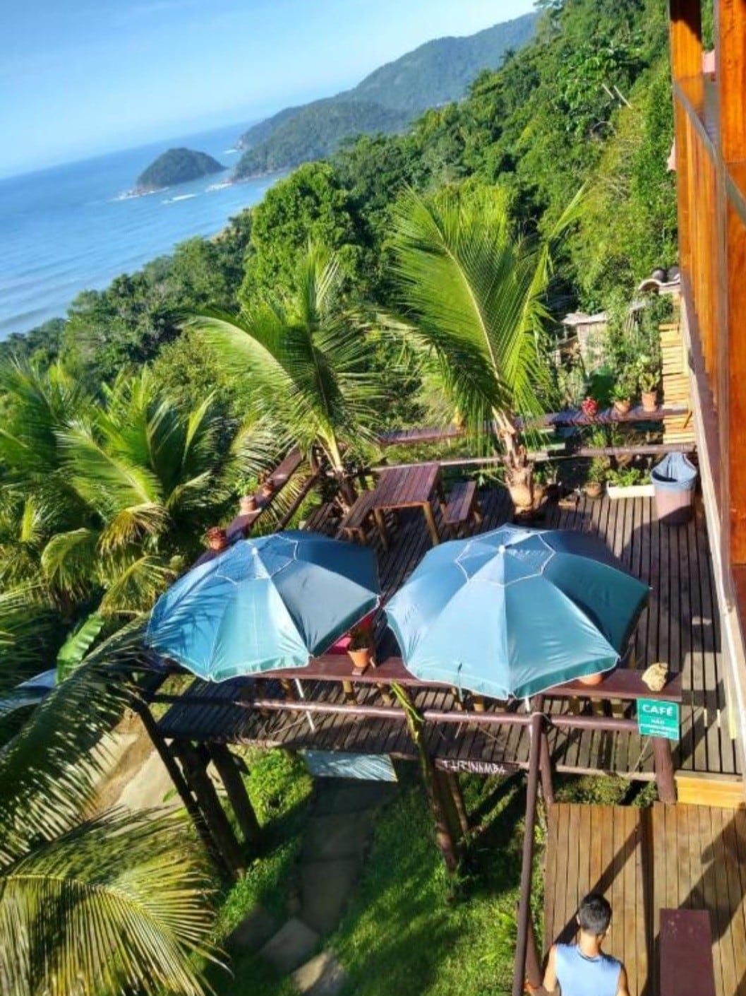 The outdoor deck features multiple seating areas shaded by large umbrellas, overlooking a stunning view of the ocean and lush greenery. Tropical plants frame the space, enhancing the connection to nature, while the wooden flooring adds a rustic touch to the environment.