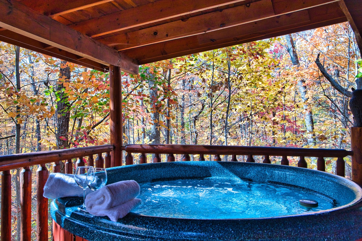 A private hot tub sits on a wooden deck surrounded by vibrant autumn foliage, providing a serene forest view. Soft towels and glassware are placed neatly beside the hot tub, inviting relaxation in a peaceful outdoor setting.