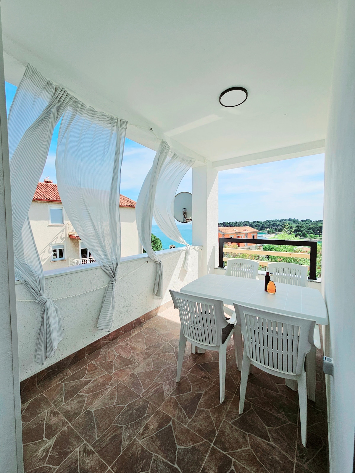 A covered balcony is presented, featuring a white table set with four matching chairs. Sheer curtains gently flow with the breeze, framing the space. A serene view of greenery and rooftops is visible in the background, contributing to a relaxing atmosphere.